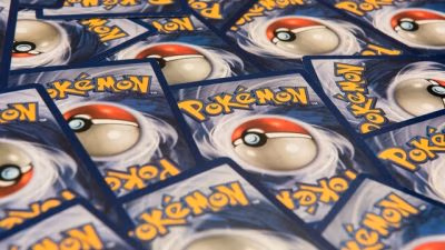 A scattered arrangement of Pokémon trading cards shown face-down, featuring the classic blue card backs with a central Poké Ball design and yellow “Pokémon” lettering. The cards overlap at different angles, filling the frame in a repeating pattern.