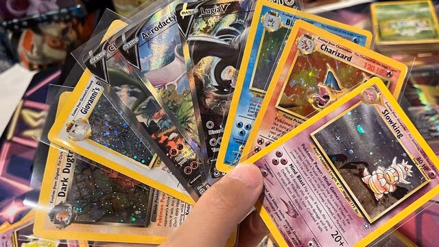 A hand holding a fan of Pokémon trading cards, including both modern and vintage styles. Visible cards include holographic and full-art designs such as Charizard, Lugia V, Aerodactyl, Dark Dugtrio, and Slowking. The cards vary in color and layout, with some featuring classic yellow borders and others modern full-art styles, all slightly overlapping in a spread.