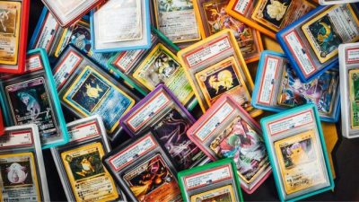 A large assortment of graded Pokémon trading cards in colorful PSA slabs is spread across a surface. The cards overlap in a dense arrangement, showing a mix of vintage and modern Pokémon artwork, including holographic and full-art cards. The slabs feature PSA labels at the top and protective cases in various colors such as blue, red, pink, and clear. The image highlights the variety and collectibility of professionally graded Pokémon cards.